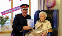 An elderly woman sits in a chair holding a card, next to a man in a formal uniform. Birthday decorations and a balloon surround them.
