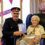 An elderly woman sits in a chair holding a card, next to a man in a formal uniform. Birthday decorations and a balloon surround them.