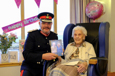 An elderly woman sits in a chair holding a card, next to a man in a formal uniform. Birthday decorations and a balloon surround them.