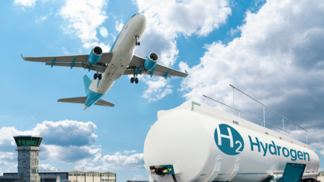 Airplane flying over an airport near a large hydrogen fuel tank marked with "H₂ Hydrogen" under a blue sky with clouds.