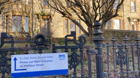 Sign for St. Olaf Street Dental Clinic advises using the main entrance on Harbour Street. Ornate black metal fence and building in the background.
