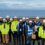 A group of people wearing hard hats and reflective jackets stand together on a coastal construction site, with the sea in the background.