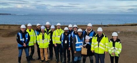 A group of people wearing hard hats and reflective jackets stand together on a coastal construction site, with the sea in the background.