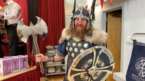 Person dressed in a Viking costume with helmet, shield, and axe stands in a room with red curtains and various items.
