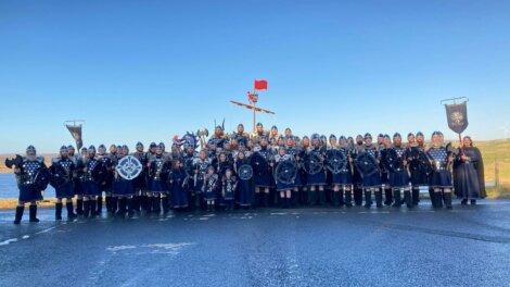 A large group of people dressed in Viking-themed costumes, holding shields and weapons, pose outdoors on a road with a clear blue sky in the background.
