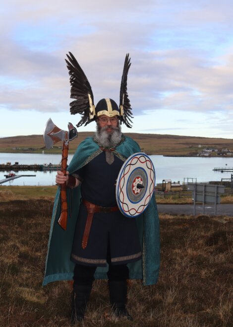 Person dressed in Viking attire with winged helmet, holding an axe and shield, standing outdoors near water and hills.