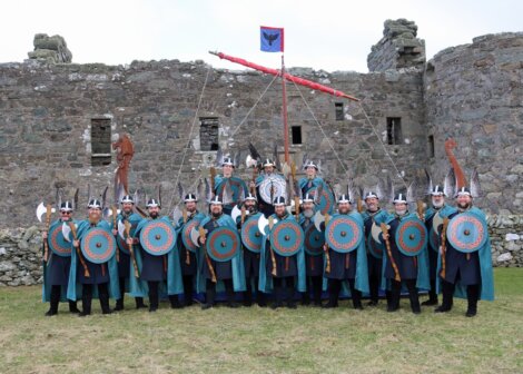 A group of people in Viking attire stand in front of an old stone structure, holding shields and axes. A flag with a bird emblem is raised behind them.