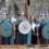 Three people dressed as Vikings with shields and axes stand next to a smiling woman in modern clothes, in front of a stone building.