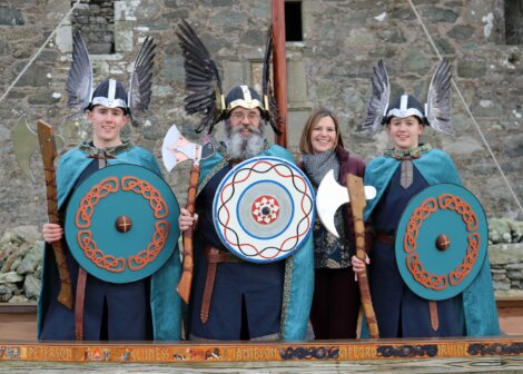 Three people dressed as Vikings with shields and axes stand next to a smiling woman in modern clothes, in front of a stone building.