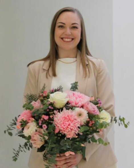 A woman smiles while holding a large bouquet of pink and white flowers, wearing a beige blazer over a white top.