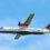 Twin-engine Loganair airplane with a red and black tartan tail livery flying against a blue sky.