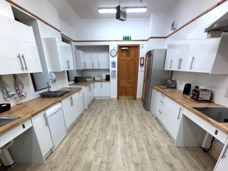 Spacious kitchen with wooden flooring, white cabinets, and matching countertops. Equipped with a sink, dishwasher, fridge, microwave, and a wall clock above a wooden door.