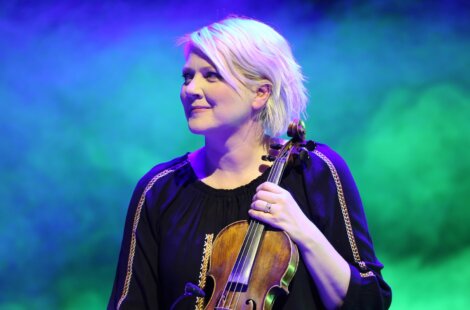 A person with short blond hair holds a violin on stage, wearing a black top with decorative trim, set against a colorful, blurred background.