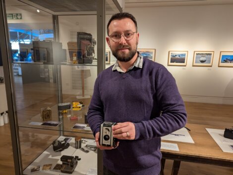 A man holding a vintage camera stands in a gallery with photos and cameras on display in the background.