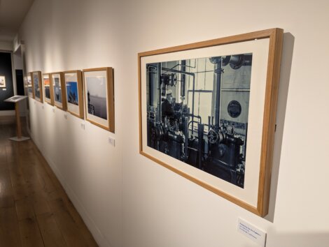 Framed photographs in a gallery setting are displayed along a white wall above a wooden floor.
