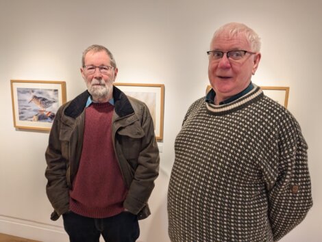 Two older men standing in an art gallery with framed photos on the wall behind them.