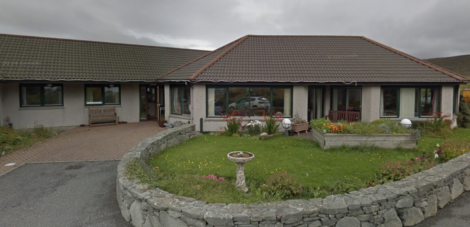 Single-story house with brown roof, white walls, and large front windows. Stone wall surrounds a garden with flowers and a birdbath. Cloudy sky overhead.