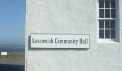 White building with a sign reading "Levenwick Community Hall" on the wall. A window is visible, and the background shows a blue sky and distant land across the water.