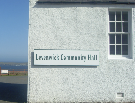 White building with a sign reading "Levenwick Community Hall" on the wall. A window is visible, and the background shows a blue sky and distant land across the water.