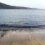 Calm sea waves reaching a sandy shore with a rocky coastline and cliffs in the background under a clear sky.