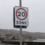 A road sign indicating a 20 mph speed limit zone on a cloudy day, with houses, hills, and a body of water in the background.