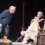 An older man in yellow boots leans forward, speaking to an older woman seated with a blanket and cane, on a stage set designed to look like a home.