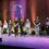 A group of eight young people stands on stage under stage lights, with a Shetland Youth Music Festival 2025 sign projected on the wood-paneled wall behind them.