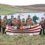 A group of people dressed as Vikings stand behind a model Viking ship with shields and axes, beside a grassy lake in a rural landscape.