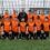 A men's football team wearing orange and black kits poses for a group photo on an artificial turf pitch in front of a wire fence.