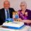 An elderly woman wearing a sash and tiara sits beside a man in a suit, celebrating her 100th birthday with a decorated cake and a card featuring a couple.