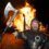 A man in Viking costume holds an axe and smiles in front of a large bonfire shaped like a Viking ship at night.
