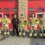 Seven firefighters in uniform, holding helmets and standing in a row outside a building with red garage doors. One person in black clothing stands among them.