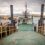 A car ferry with its ramp lowered is docked at a pier, ready to load vehicles. Water and a rocky shoreline are visible in the background under a cloudy sky.