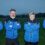 Four adults in matching blue and black team jackets stand side by side with arms crossed on a grassy field, facing the camera.