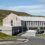 A modern, two-story building with a sloped metal roof, large windows, and a small parking area, set against grassy hills under a partly cloudy sky.