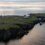 Aerial view of a grassy coastal island with a lighthouse, buildings, and cliffs, surrounded by calm sea under a cloudy sky at sunset.
