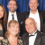 Six adults dressed in formal attire pose and smile for a group photo in front of a blue curtain backdrop.