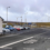 A parking lot with several cars next to a small building, adjacent to a road marked "STOP," with wind turbines visible in the background.