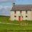 A two-story, gray stone house with a red door stands in a grassy rural setting, surrounded by a fence and open fields under a partly cloudy sky.