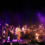 A large band performs on stage with various instruments including guitars, keyboard, violin, and drums, illuminated by colored stage lights and surrounded by smoke effects.