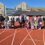 A group of adults and children stand on an outdoor running track in front of a modern building on a clear, sunny day.