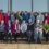 A group of around thirty people, mostly women, pose outside in two rows. Many are wearing colorful knit hats and sweaters. They are in front of a stone wall and glass building.