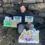 A person sits outdoors against a stone wall, holding two illustrated children's books and flanked by two large, colorful cutouts of cartoon characters.