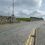 A paved road with double yellow lines runs uphill past stone walls, with a few cars parked on the right and houses visible in the background under a cloudy sky.