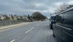 A wide, empty road with parked vans on the right, stone buildings on the left, overcast sky, and a few distant pedestrians and cars.