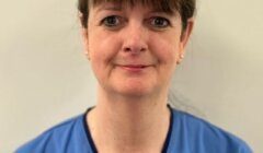 A woman in blue NHS Scotland scrubs stands against a plain light-colored background, looking at the camera with a neutral expression.
