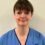 A woman in blue NHS Scotland scrubs stands against a plain light-colored background, looking at the camera with a neutral expression.
