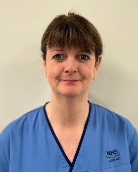 A woman in blue NHS Scotland scrubs stands against a plain light-colored background, looking at the camera with a neutral expression.