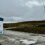 A sign reading “Welcome to Viking Energy Wind Farm” stands at the entrance, with wind turbines visible on a grassy, overcast landscape in the background.