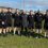 A group of ten men in black sportswear stand in a line on a grass field, posing for a photo on a sunny day.